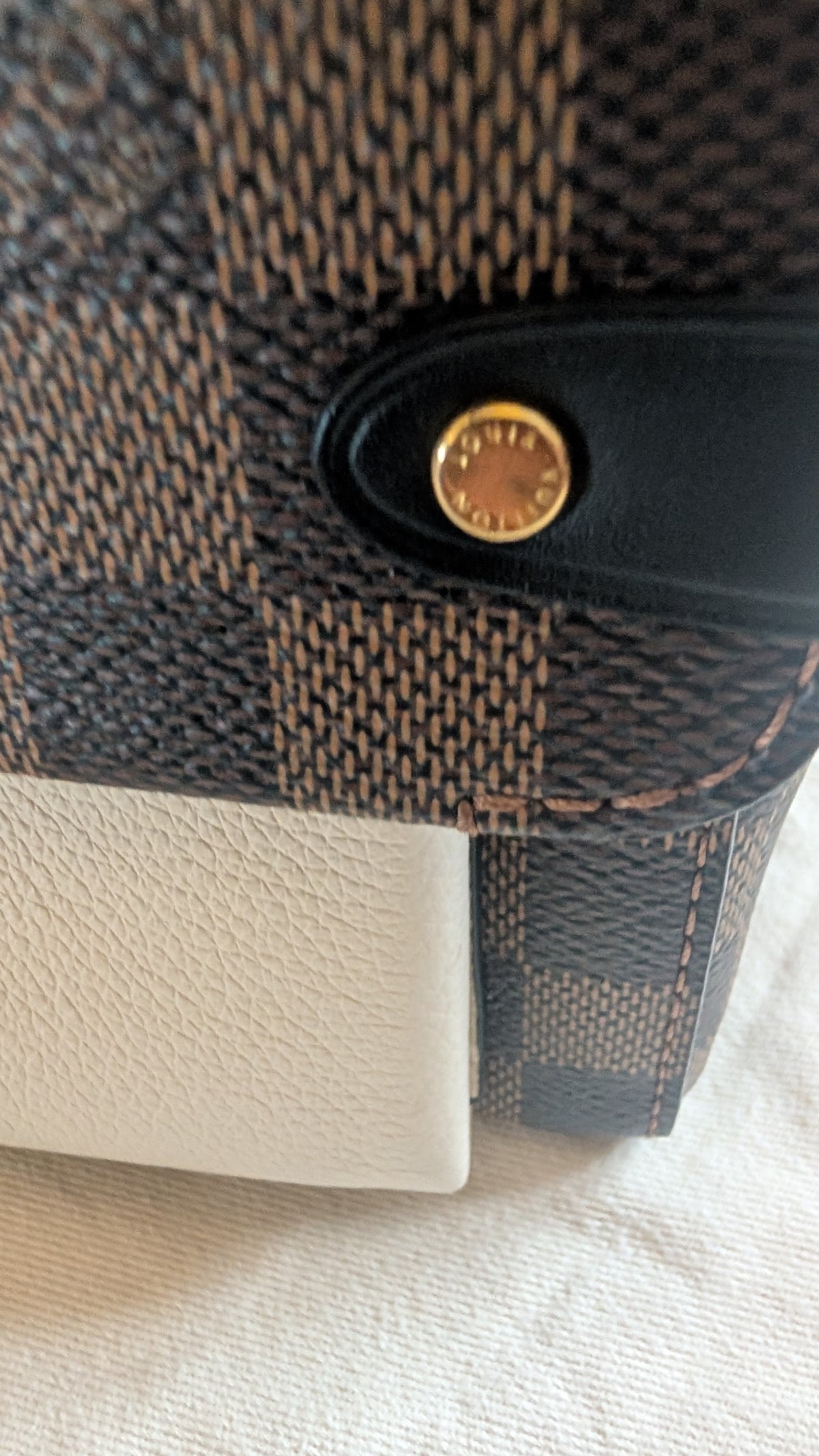 Close-up of a Louis Vuitton handbag corner showing the dark brown and beige monogram canvas, a black leather tab with a gold-tone circular logo, and a white textured leather section on a light fabric background. Pre-Owned Louis Vuitton Vavin PM Damier Ebene / Crème – N40113