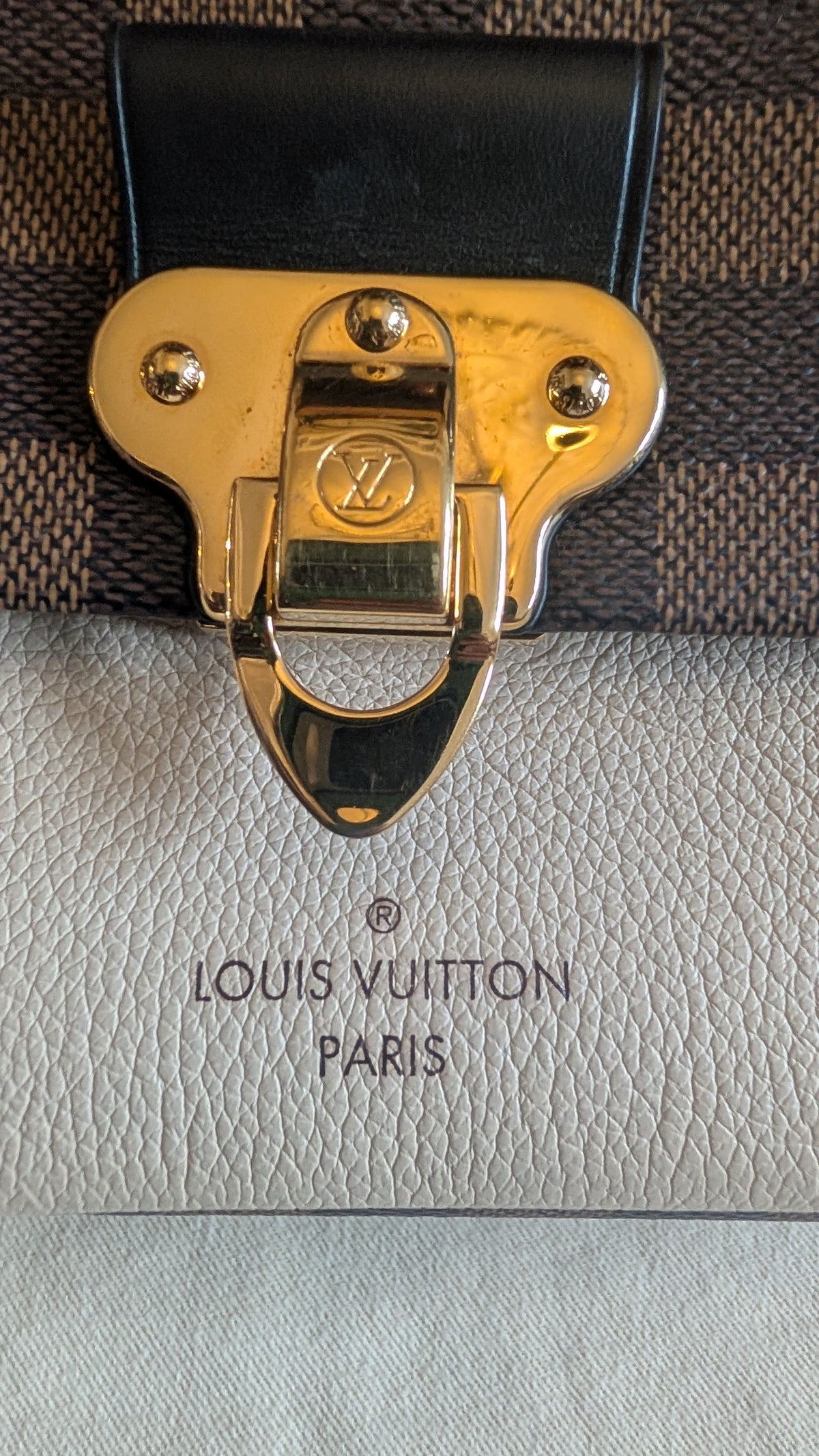 Close-up of a Louis Vuitton bag showing a gold-tone metal clasp on a dark brown and beige patterned canvas, with a light beige textured leather section below embossed with 'LOUIS VUITTON PARIS'. Pre-Owned Louis Vuitton Vavin PM Damier Ebene / Crème – N40113