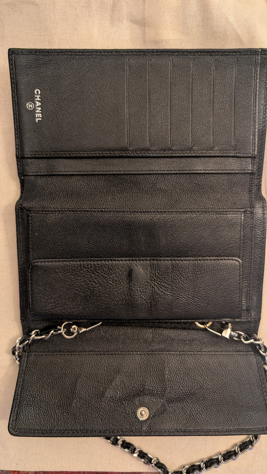 An open black leather wallet or clutch with multiple card slots, a chain strap, and a silver-tone snap closure, photographed against a light beige fabric background. The brand name CHANEL is visible on the interior. Pre-Owned Chanel Vintage Black Caviar Leather Wallet with Added Chain