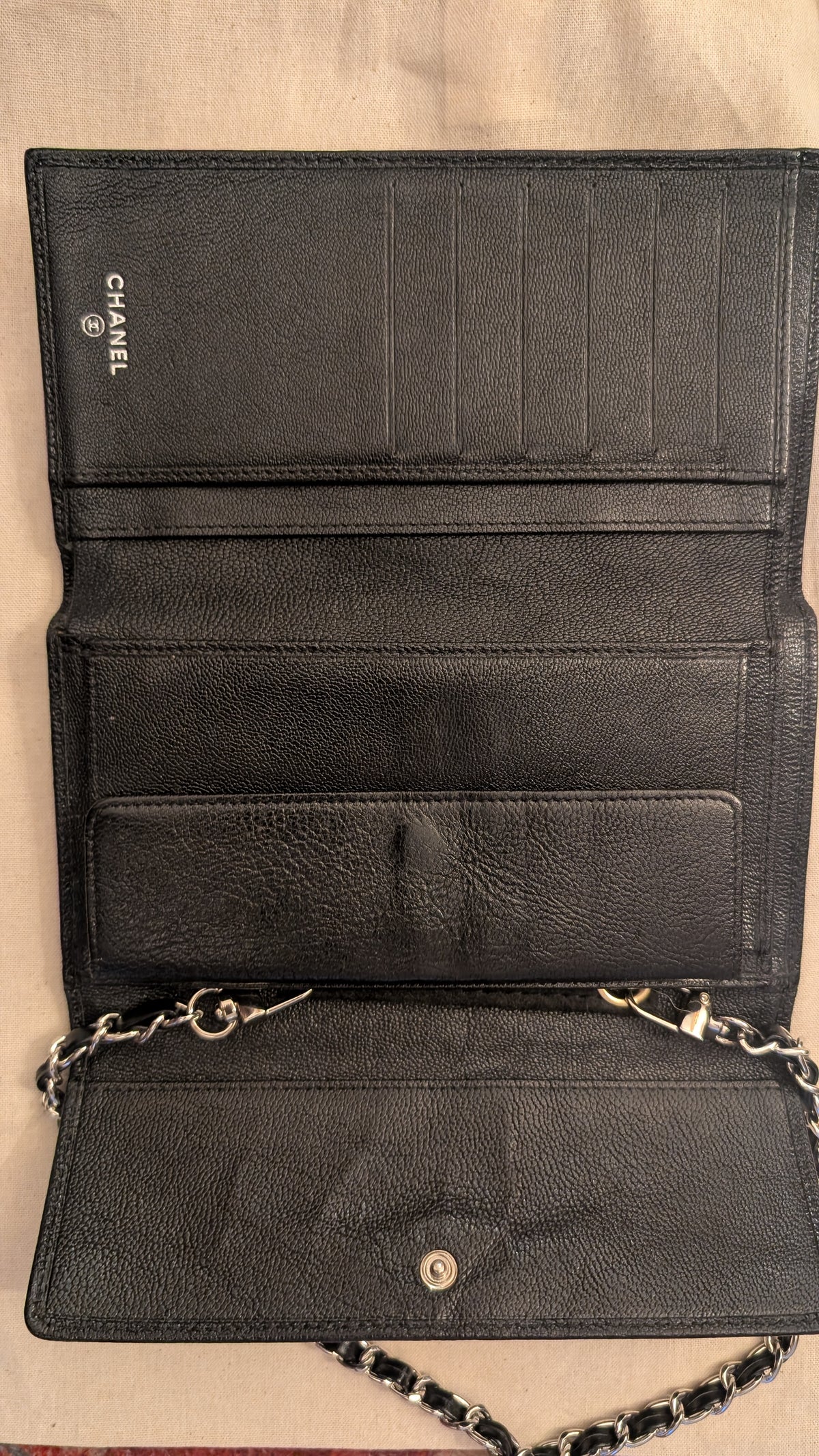 An open black leather wallet or clutch with multiple card slots, a chain strap, and a silver-tone snap closure, photographed against a light beige fabric background. The brand name CHANEL is visible on the interior. Pre-Owned Chanel Vintage Black Caviar Leather Wallet with Added Chain