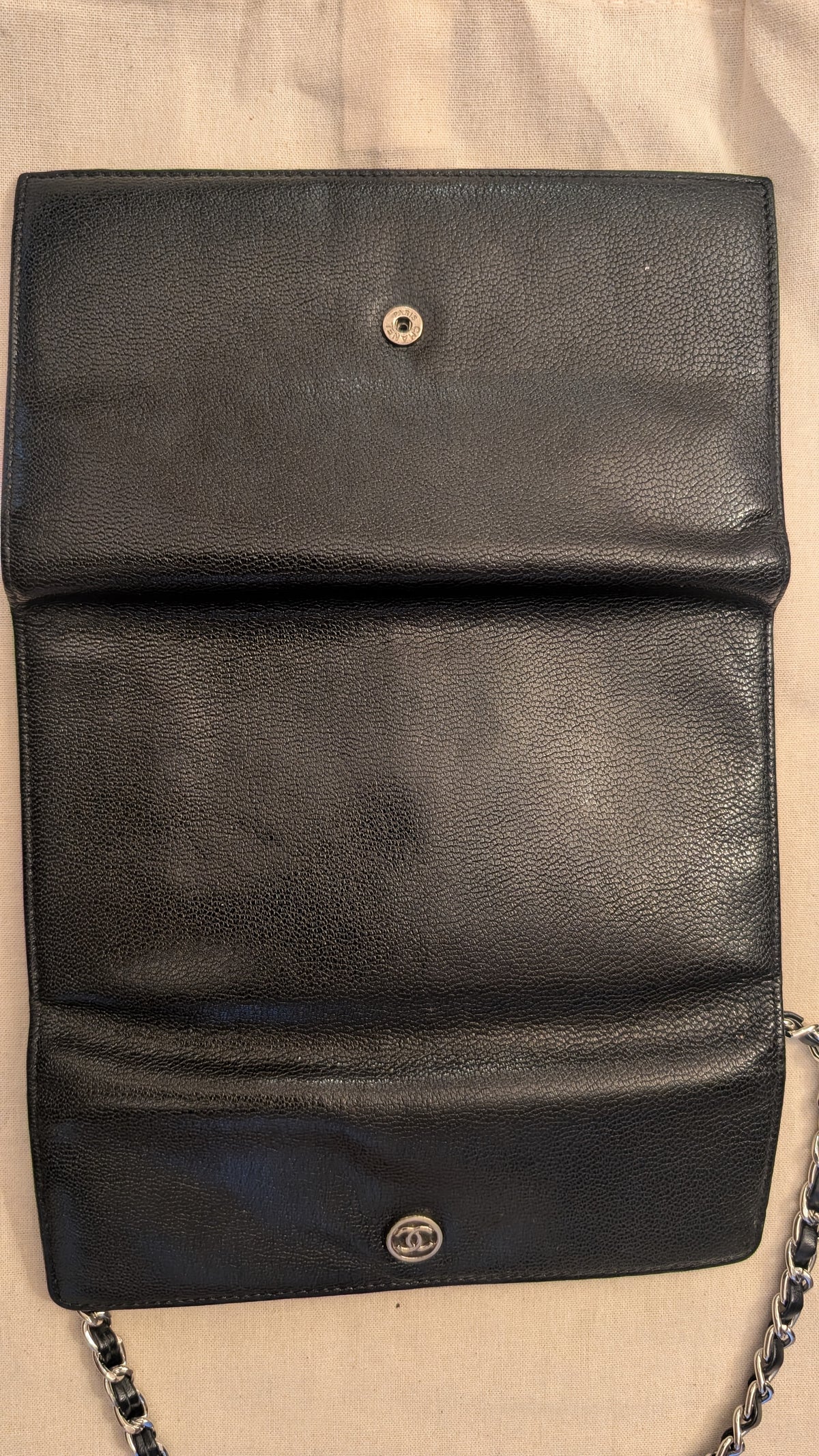 A black pebbled leather shoulder bag with a silver-tone metal snap button and chain strap, laid flat on a beige fabric background. Pre-Owned Chanel Vintage Black Caviar Leather Wallet with Added Chain