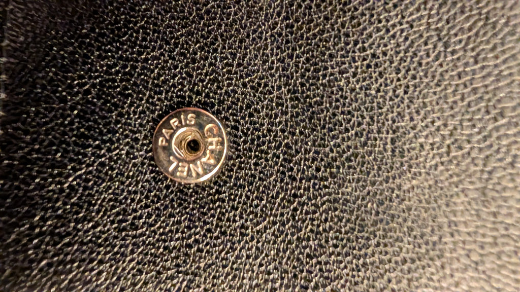 Close-up of a black pebbled leather surface with a round metal Chanel snap button showing the engraved brand  Pre-Owned Chanel Vintage Black Caviar Leather Wallet with Added Chainname.