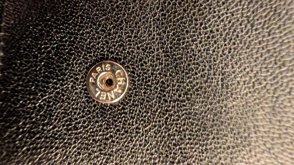 Close-up of a black pebbled leather surface with a round metal Chanel snap button showing the engraved brand  Pre-Owned Chanel Vintage Black Caviar Leather Wallet with Added Chainname.