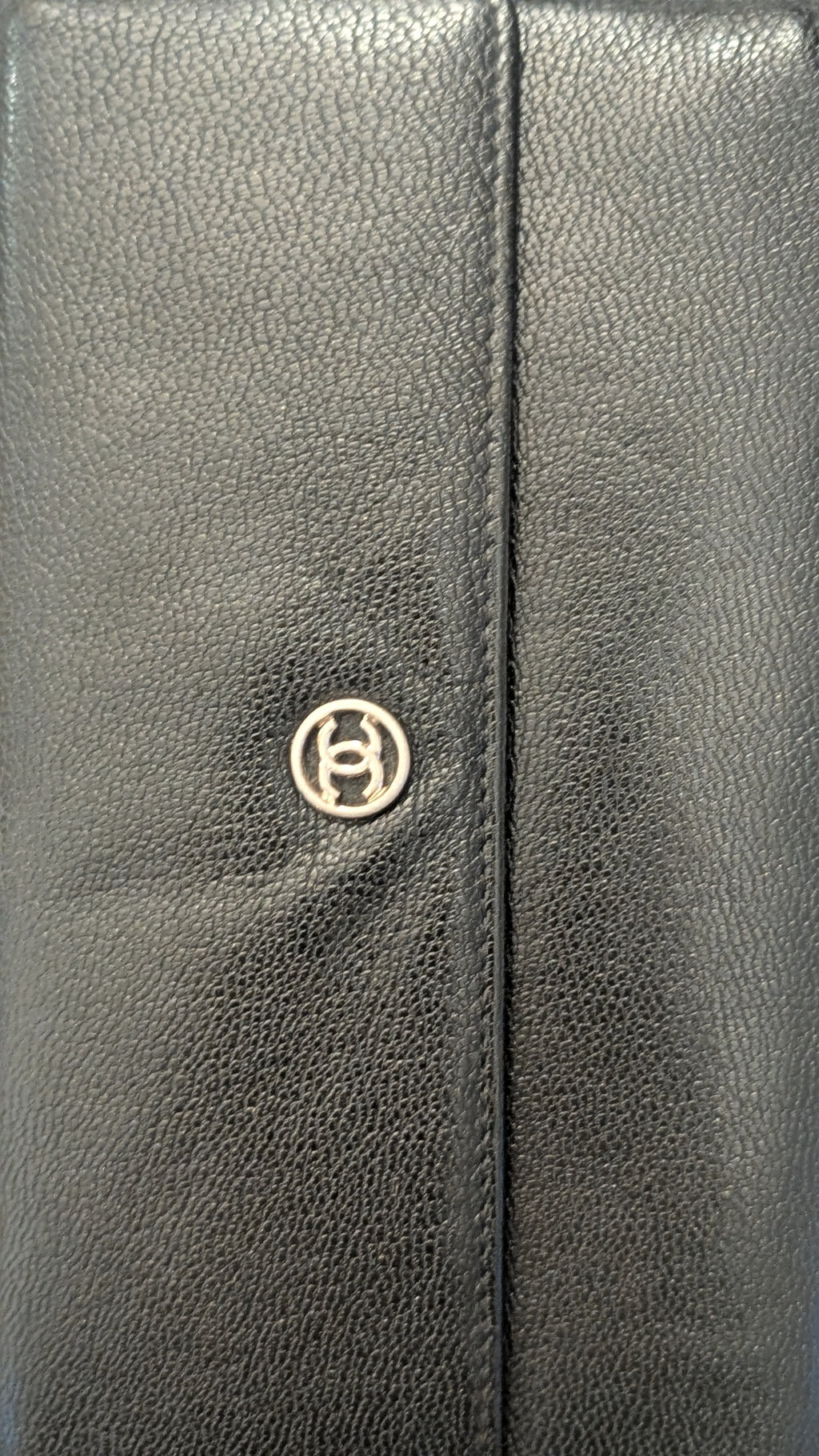 Close-up of a black pebbled leather item, likely a wallet or clutch, featuring a vertical center seam and a small circular metallic logo emblem. Pre-Owned Chanel Vintage Black Caviar Leather Wallet with Added Chain