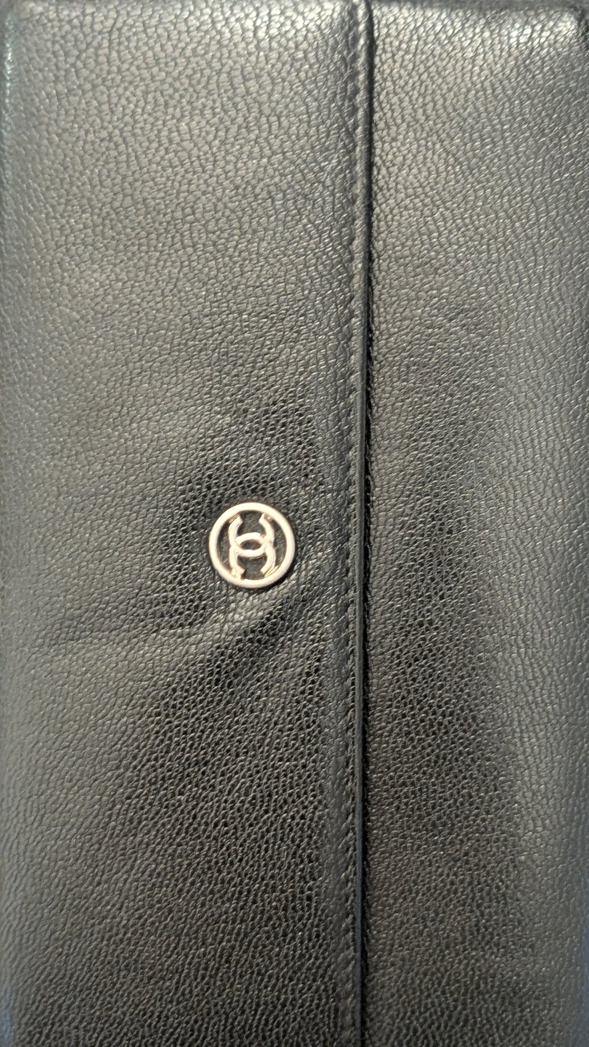 Close-up of a black pebbled leather item, likely a wallet or clutch, featuring a vertical center seam and a small circular metallic logo emblem. Pre-Owned Chanel Vintage Black Caviar Leather Wallet with Added Chain