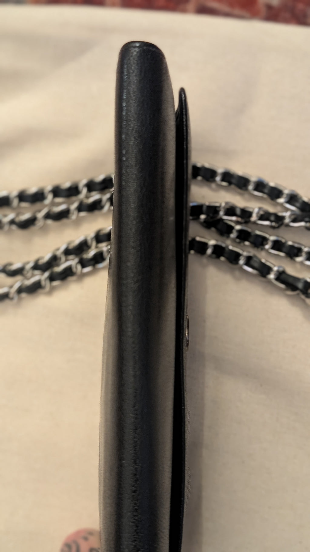A close-up side view of a black leather wallet or card holder with a black and silver chain strap, resting on a light beige fabric surface. Pre-Owned Chanel Vintage Black Caviar Leather Wallet with Added Chain