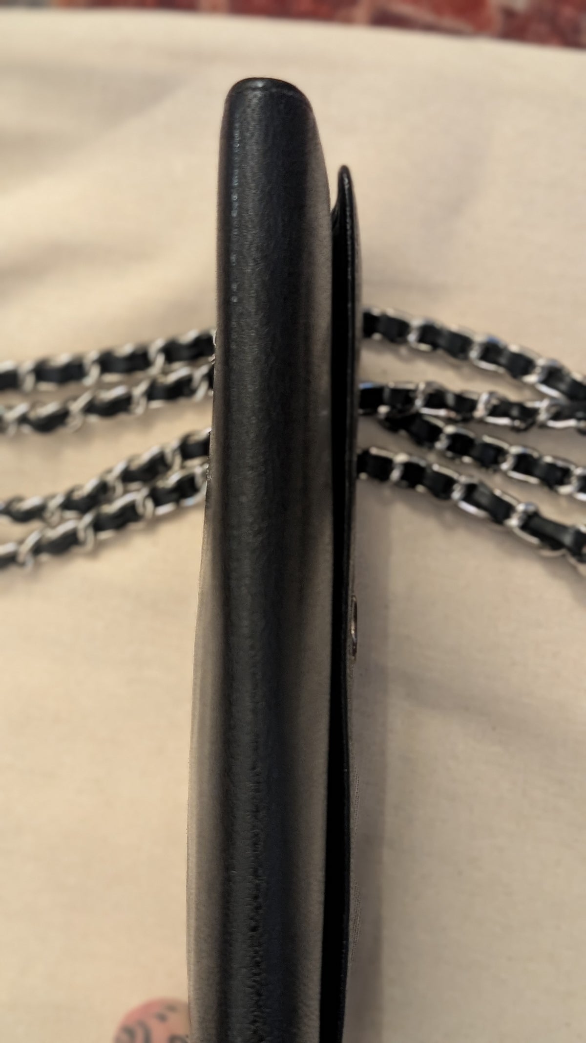 A close-up side view of a black leather wallet or card holder with a black and silver chain strap, resting on a light beige fabric surface. Pre-Owned Chanel Vintage Black Caviar Leather Wallet with Added Chain