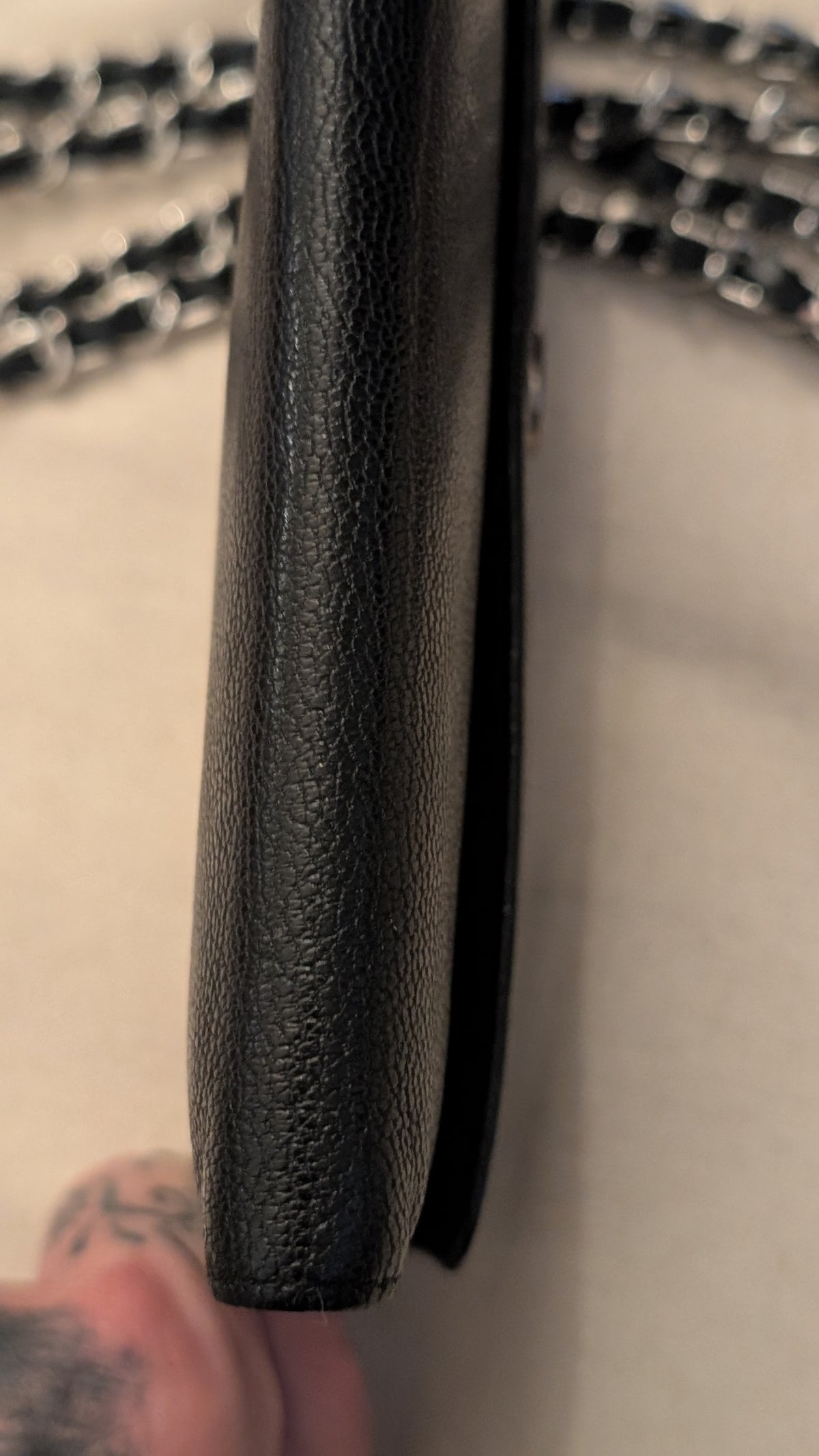 Close-up of a black pebbled leather wallet or clutch shown vertically, with a silver chain strap visible in the blurred background and a hand holding the bottom edge. Pre-Owned Chanel Vintage Black Caviar Leather Wallet with Added Chain