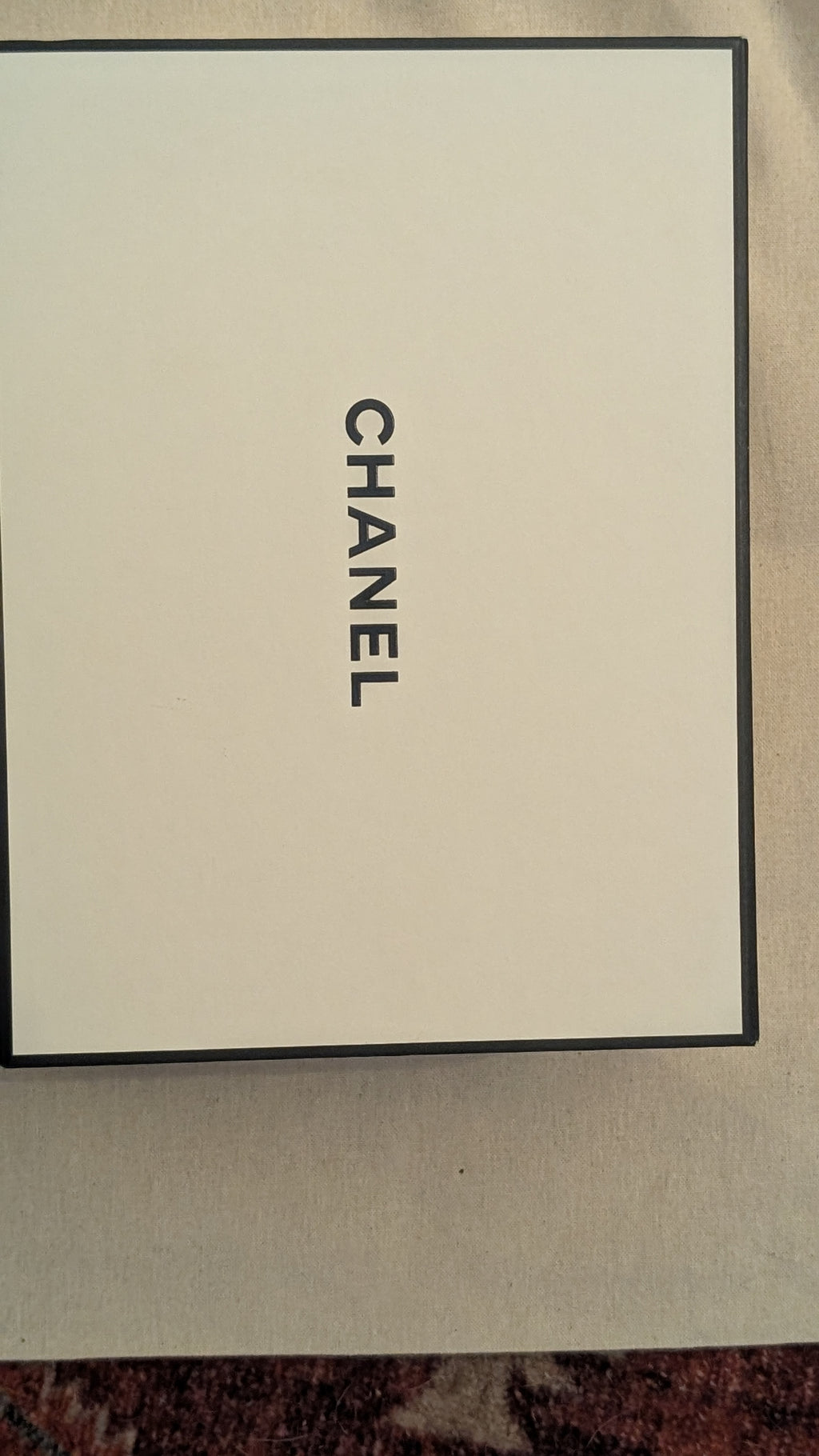 A Chanel-branded cream-colored box with a black border and vertical black CHANEL text, photographed from above on a neutral surface. Pre-Owned Chanel Vintage Black Caviar Leather Wallet with Added Chain