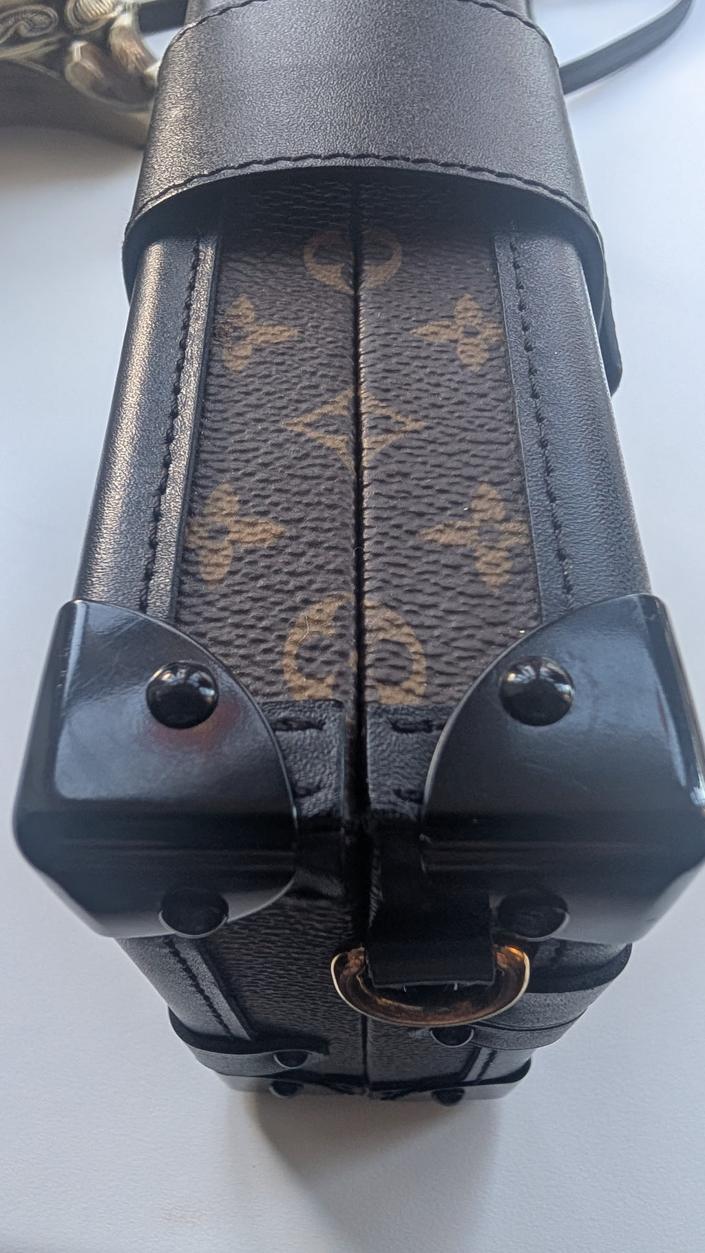Close-up of a Louis Vuitton monogram canvas and leather bag corner with black hardware and a gold-tone ring detail, photographed on a light surface. Pre-Owned Louis Vuitton Petite Malle Monogram Canvas Crossbody Bag