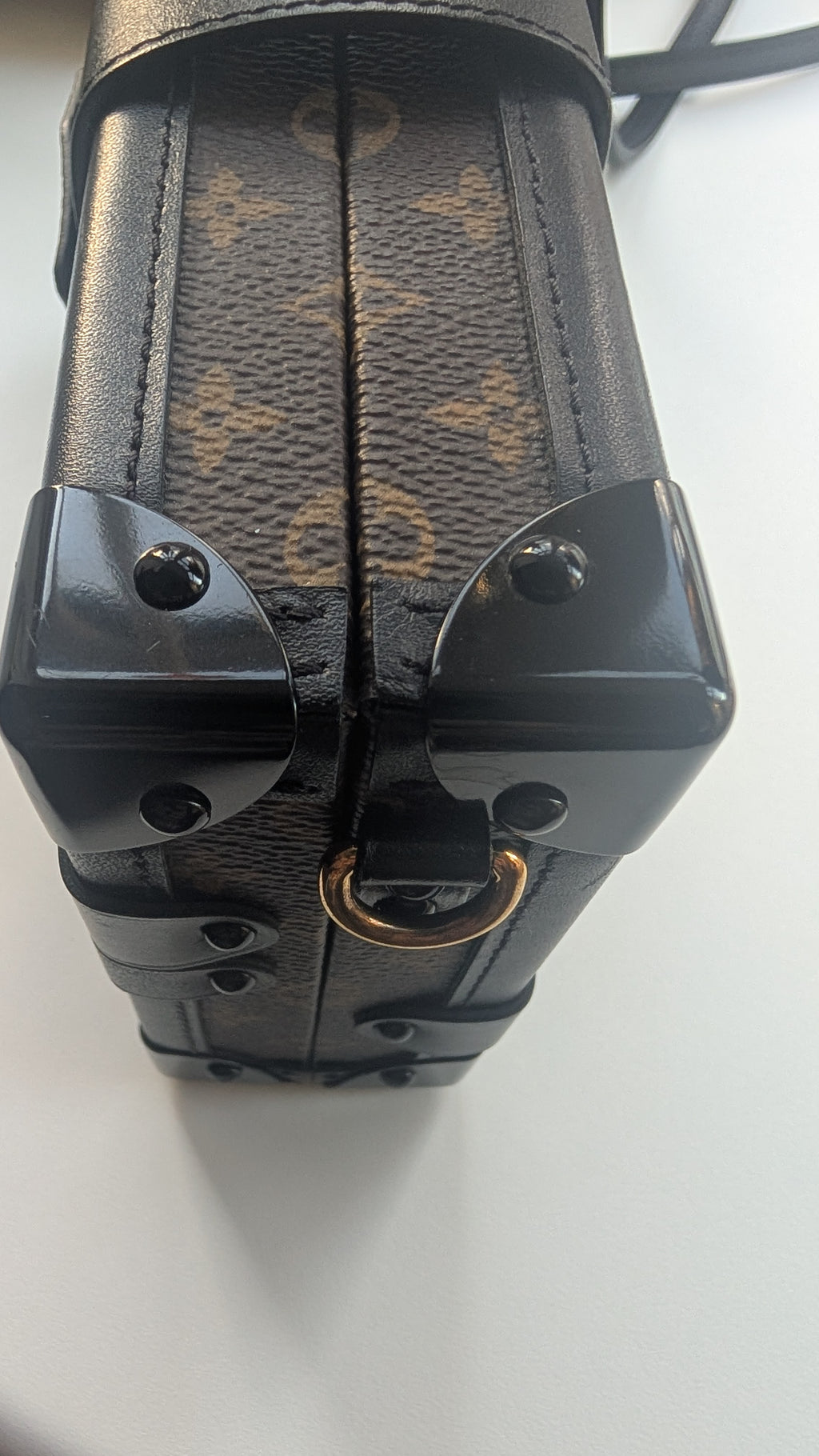Close-up of a Louis Vuitton monogram canvas and leather bag corner with black hardware and a gold-tone ring on a white surface. Pre-Owned Louis Vuitton Petite Malle Monogram Canvas Crossbody Bag