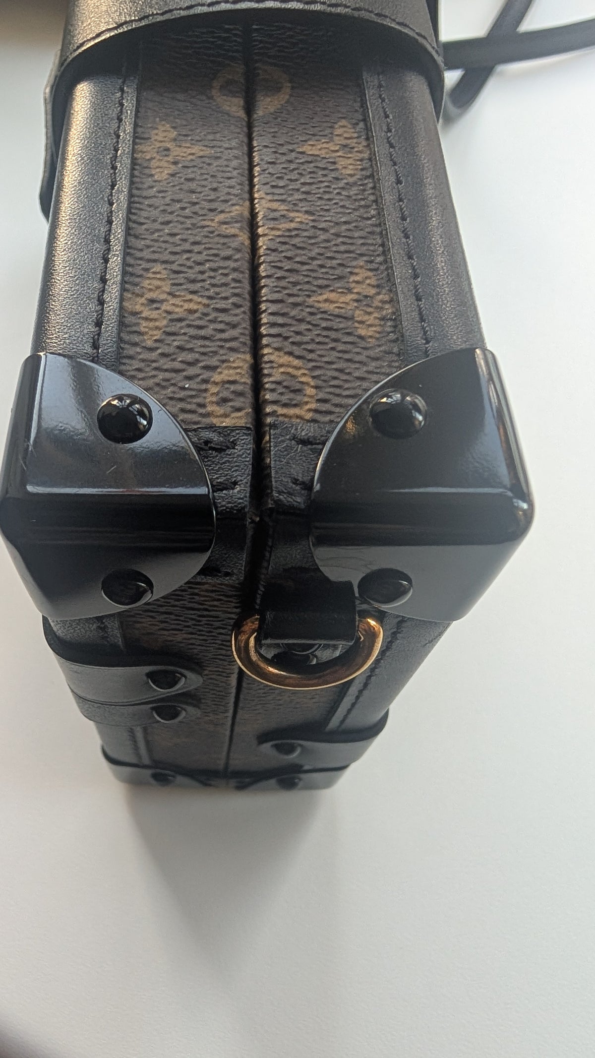 Close-up of a Louis Vuitton monogram canvas and leather bag corner with black hardware and a gold-tone ring on a white surface. Pre-Owned Louis Vuitton Petite Malle Monogram Canvas Crossbody Bag