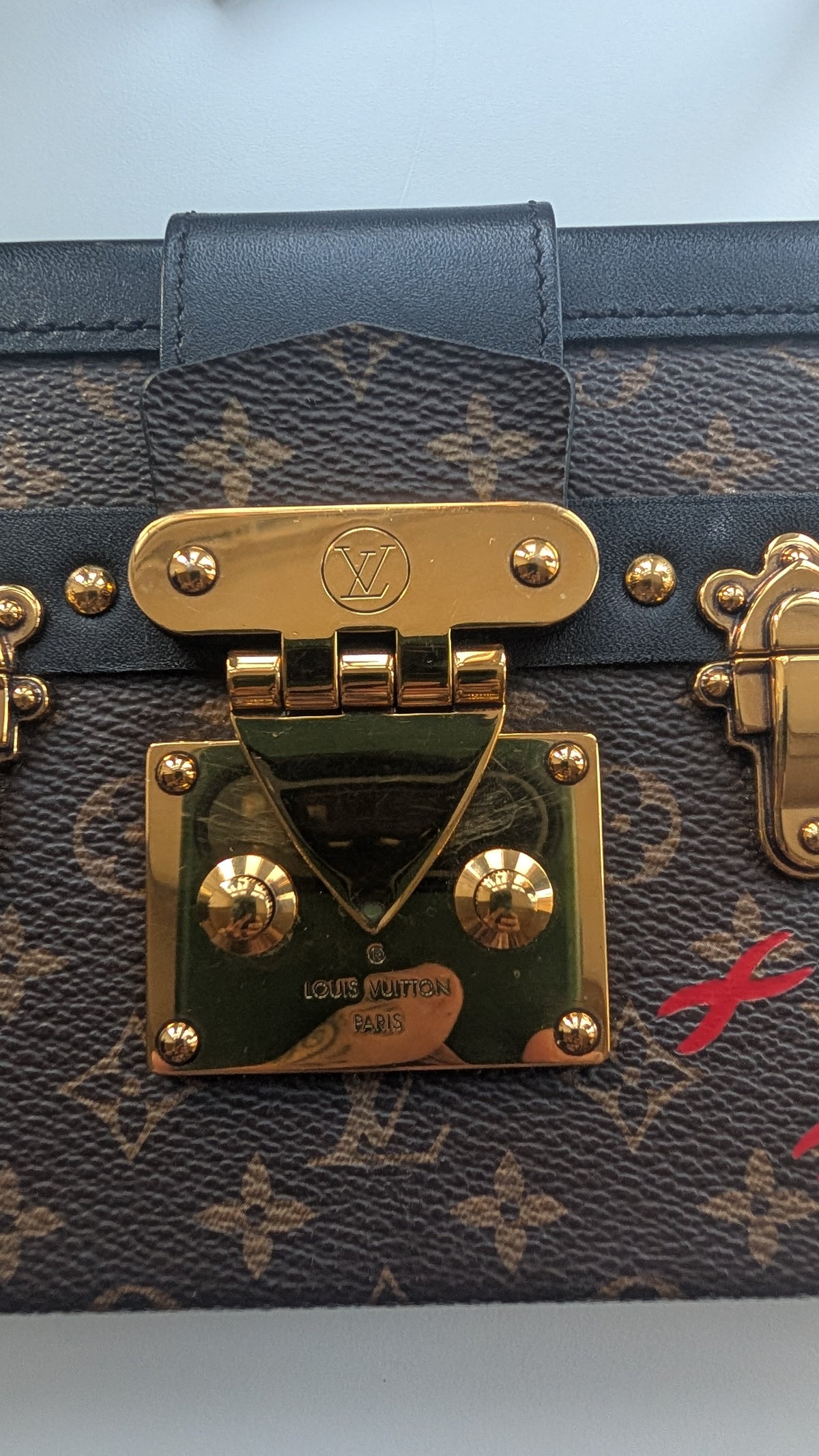 Close-up of a Louis Vuitton monogram canvas bag with a dark blue leather strap and prominent gold-tone hardware, including a central clasp with the LV logo. Pre-Owned Louis Vuitton Petite Malle Monogram Canvas Crossbody Bag