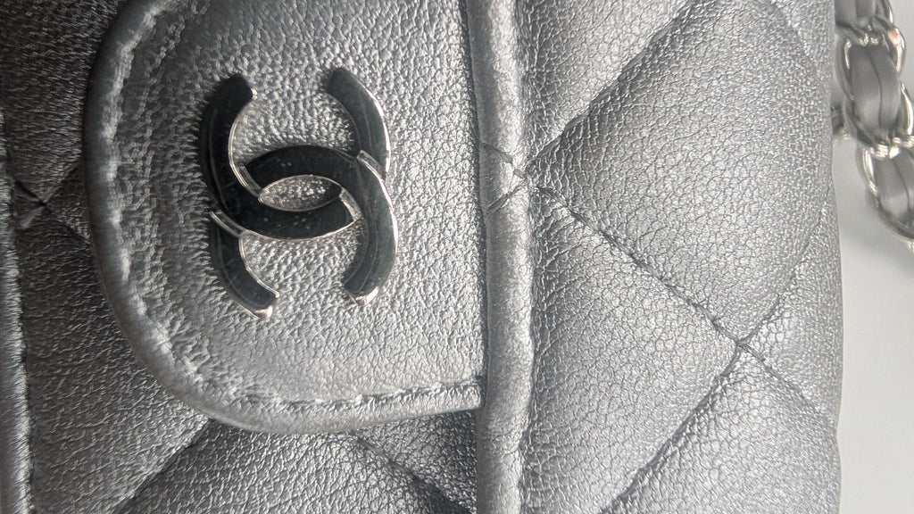 Close-up of a silver quilted leather handbag featuring a black interlocking CC logo charm on a silver-tone ring, with a silver chain strap visible on the right. Pre-Owned Chanel Metallic Silver Matelassé Mini Chain Wallet / Micro Crossbody
