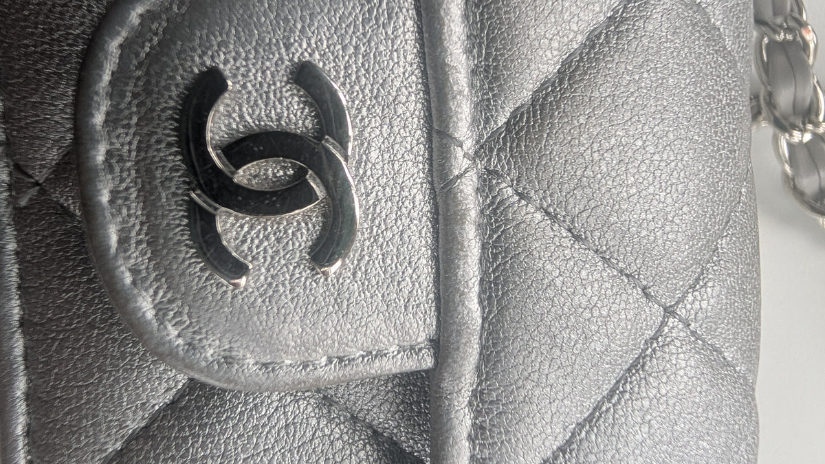 Close-up of a silver quilted leather handbag featuring a black interlocking CC logo charm on a silver-tone ring, with a silver chain strap visible on the right. Pre-Owned Chanel Metallic Silver Matelassé Mini Chain Wallet / Micro Crossbody