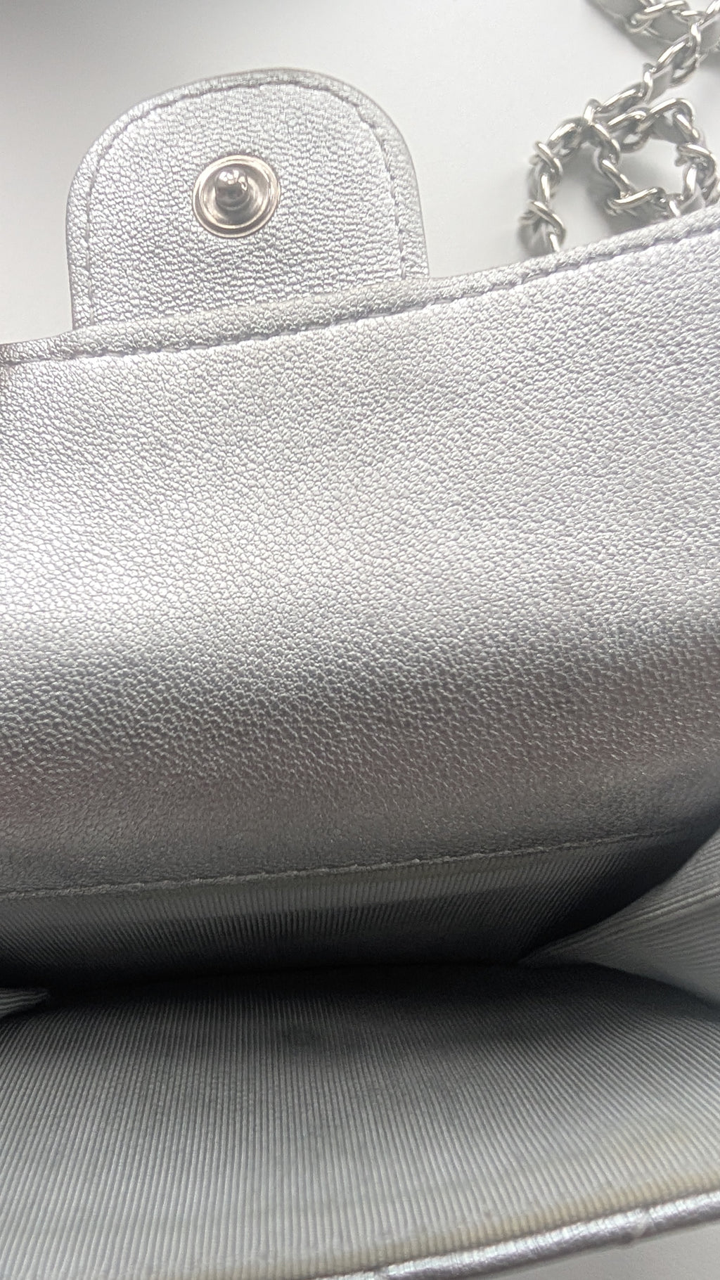 Close-up of a silver textured leather handbag showing the interior lining, a snap button closure, and part of a silver chain strap. Pre-Owned Chanel Metallic Silver Matelassé Mini Chain Wallet / Micro Crossbody
