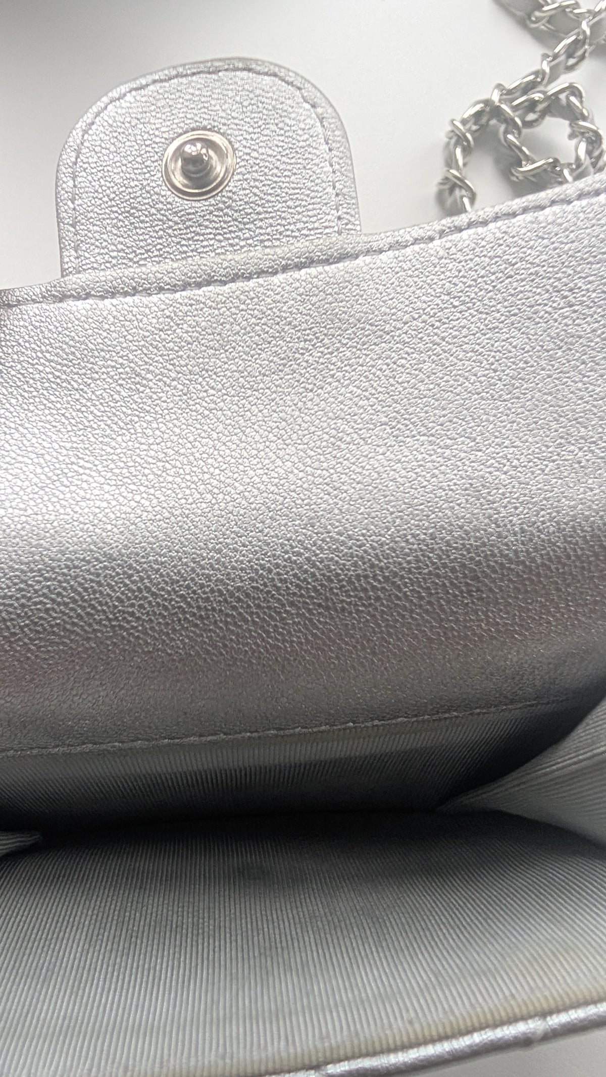 Close-up of a silver textured leather handbag showing the interior lining, a snap button closure, and part of a silver chain strap. Pre-Owned Chanel Metallic Silver Matelassé Mini Chain Wallet / Micro Crossbody