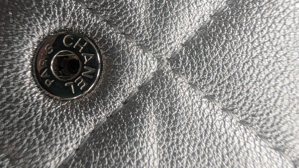 Close-up of a silver textured leather surface with a round metal snap button engraved with the brand name CHANEL. Pre-Owned Chanel Metallic Silver Matelassé Mini Chain Wallet / Micro Crossbody