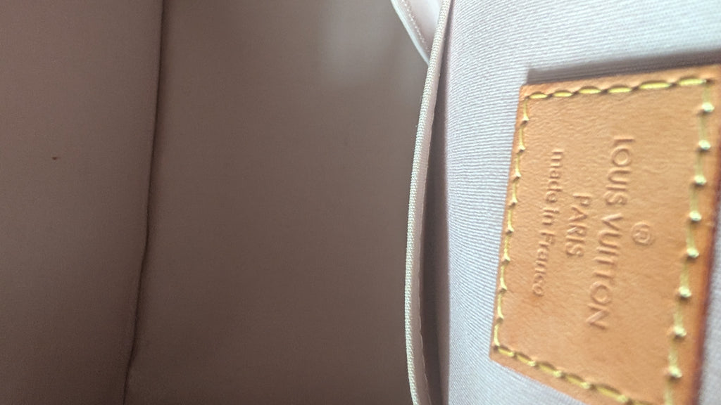 Close-up of a Louis Vuitton product showing a tan leather patch with gold-tone stitching and debossed brand text on a light gray fabric surface. Pre-Owned Louis Vuitton Alma PM Monogram Vernis – Beige Poudre
