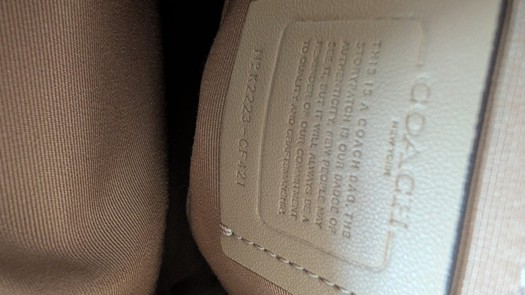 Close-up of a Coach interior label stitched onto a beige fabric surface, with the Coach name embossed on the leather patch. Pre-Owned Coach Sydney Satchel With Heart Cherry Print – CF421