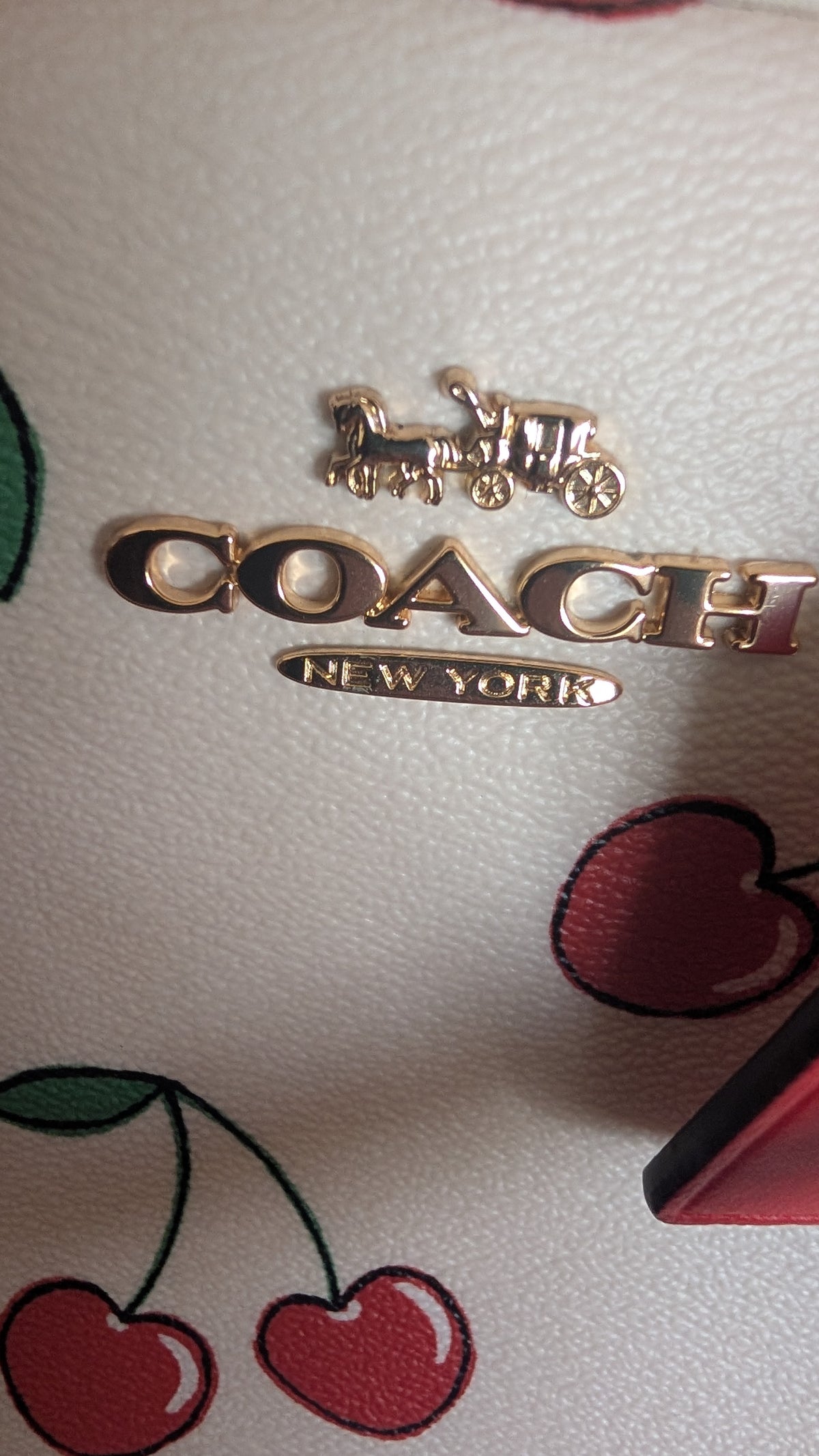 Close-up of a Coach New York metallic logo plaque on a light textured leather surface with cherry illustrations. Pre-Owned Coach Sydney Satchel With Heart Cherry Print – CF421