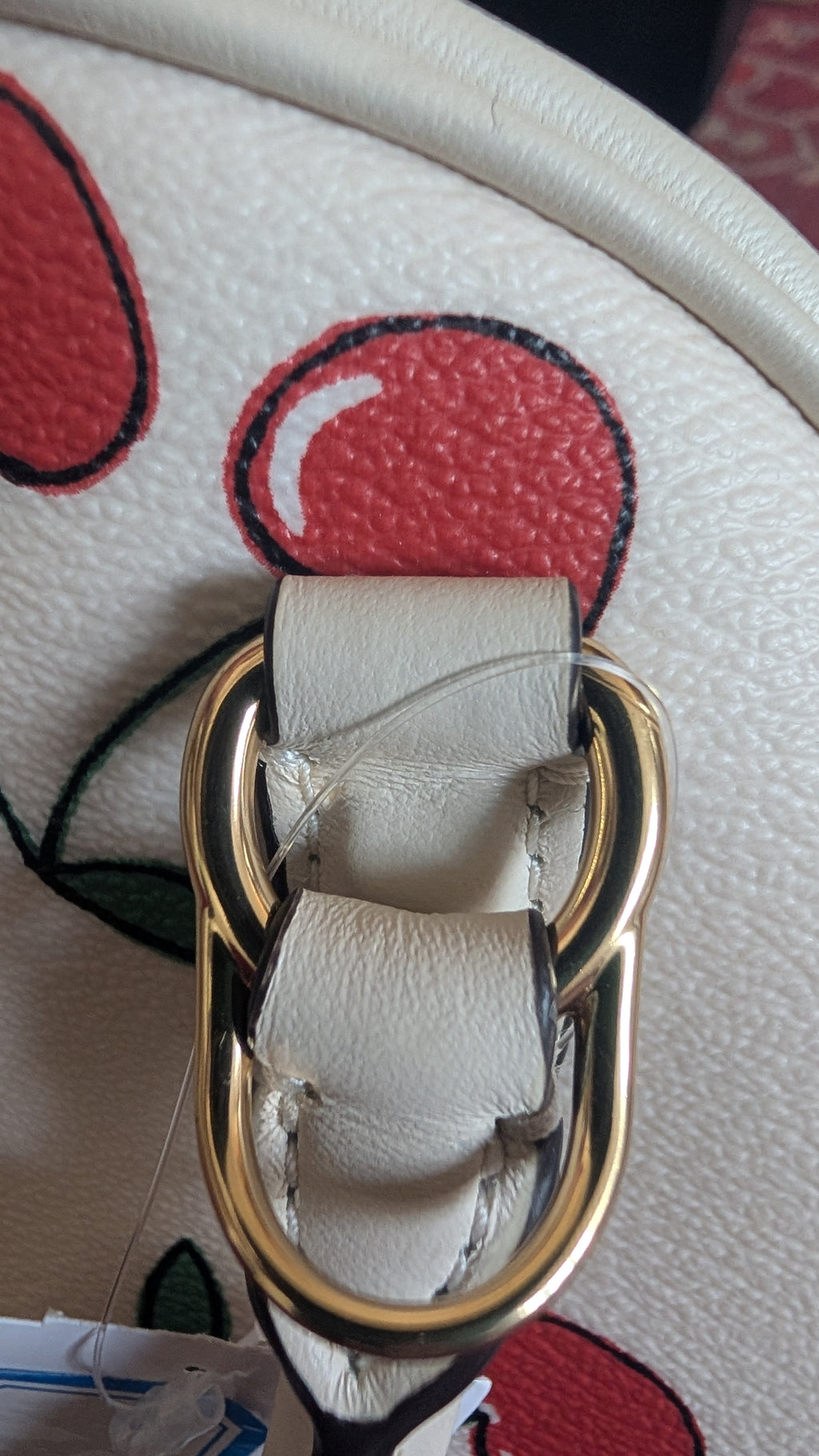 Close-up of a handbag featuring a cream-colored leather strap with a gold-tone metal O-ring and a red and black cherry print on the body. Pre-Owned Coach Sydney Satchel With Heart Cherry Print – CF421