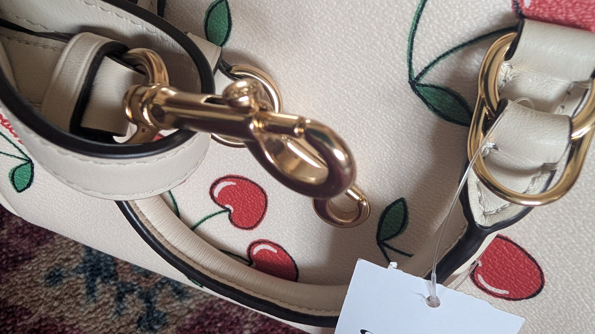 Close-up of a handbag strap and hardware, showing a cream-colored leather strap with a red and green cherry pattern, gold-tone metal rings and clasp, and a white hangtag. Pre-Owned Coach Sydney Satchel With Heart Cherry Print – CF421