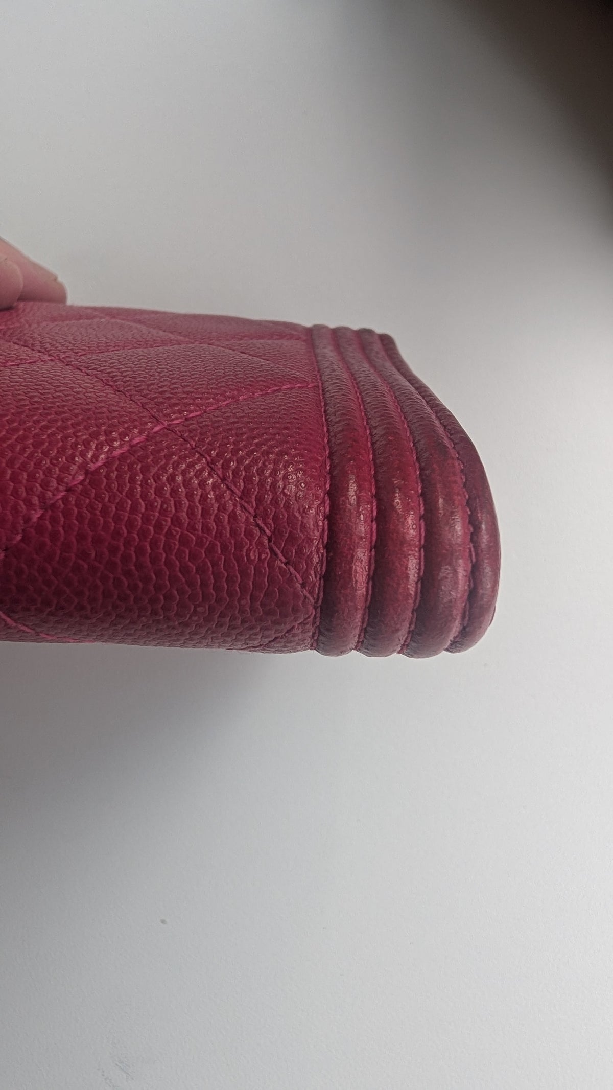 A close-up of a red pebbled leather wallet or clutch corner held by a hand, showing the textured leather and stitched edge against a plain light   Pre-Owned Chanel Boy Wallet on Chain – Fuchsia Pink Caviar Leather – Gold Hardwarebackground.