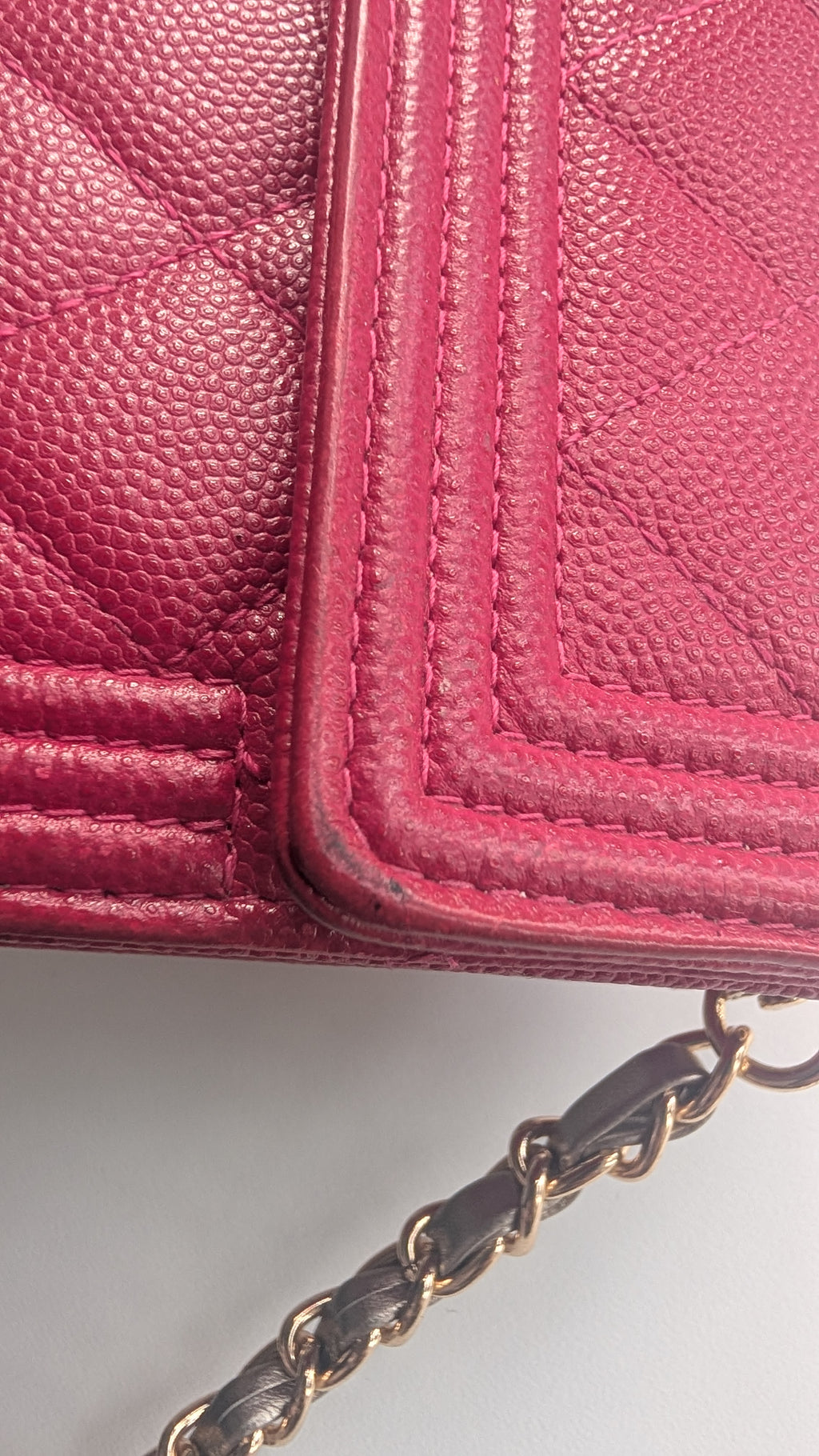 A close-up of a pink textured leather handbag corner with quilted stitching and a gold-tone chain strap visible at the bottom right. Pre-Owned Chanel Boy Wallet on Chain – Fuchsia Pink Caviar Leather – Gold Hardware