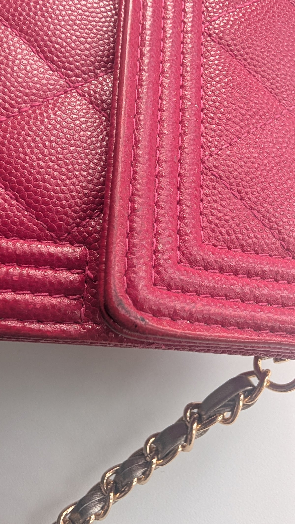 A close-up of a pink textured leather handbag corner with quilted stitching and a gold-tone chain strap visible at the bottom right. Pre-Owned Chanel Boy Wallet on Chain – Fuchsia Pink Caviar Leather – Gold Hardware