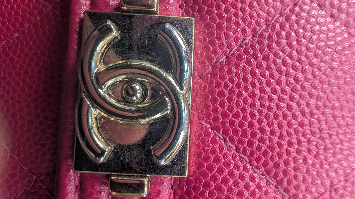 A close-up of a metallic Chanel CC turn-lock clasp with an eye motif, set against a textured magenta pink pebbled leather background. Pre-Owned Chanel Boy Wallet on Chain – Fuchsia Pink Caviar Leather – Gold Hardware