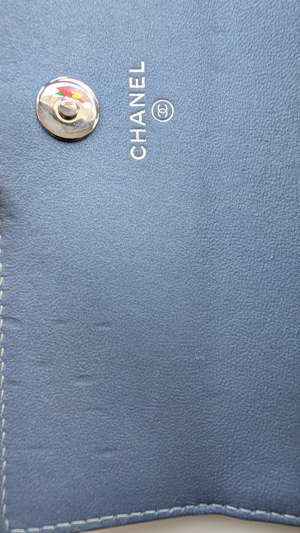 Close-up of a blue leather wallet or clutch showing the silver-tone CHANEL logo and a snap button closure. Pre-Owned Chanel Camellia Studded Wallet on Chain WOC – Blue Lambskin