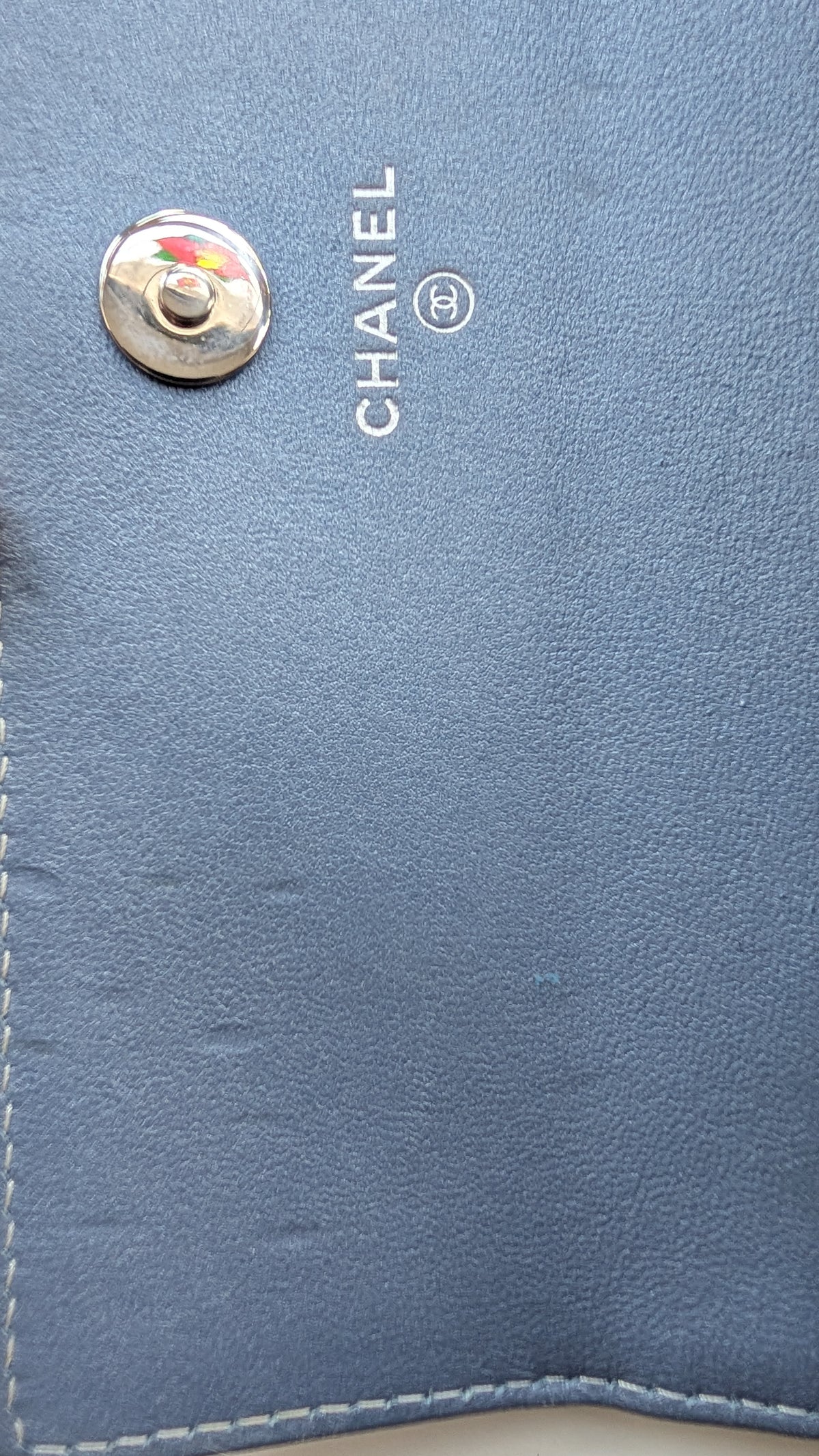 Close-up of a blue leather wallet or clutch showing the silver-tone CHANEL logo and a snap button closure. Pre-Owned Chanel Camellia Studded Wallet on Chain WOC – Blue Lambskin