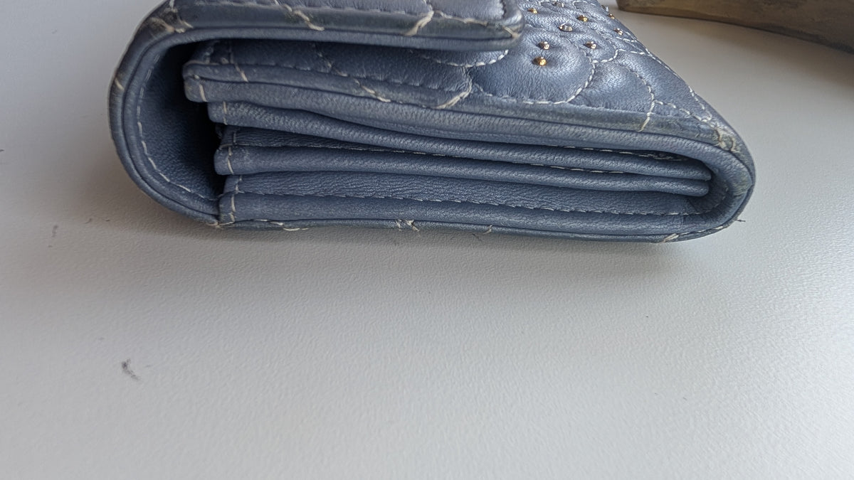 A close-up of a worn blue leather wallet or card holder showing creased, scuffed panels and visible wear on a light gray surface. Pre-Owned Chanel Camellia Studded Wallet on Chain WOC – Blue Lambskin