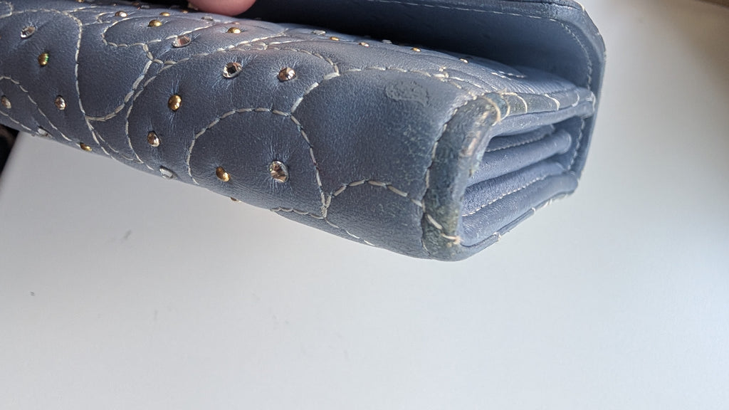 A close-up of a blue quilted leather wallet or clutch with gold-tone studs and contrast stitching, held against a plain light background. Pre-Owned Chanel Camellia Studded Wallet on Chain WOC – Blue Lambskin