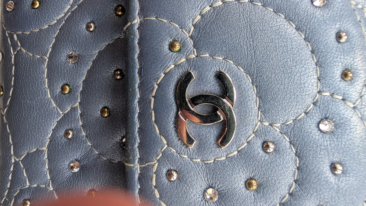 Close-up of a blue quilted leather accessory featuring a prominent interlocking CC logo in polished silver-tone metal, accented with white stitching and small metallic studs. Pre-Owned Chanel Camellia Studded Wallet on Chain WOC – Blue Lambskin