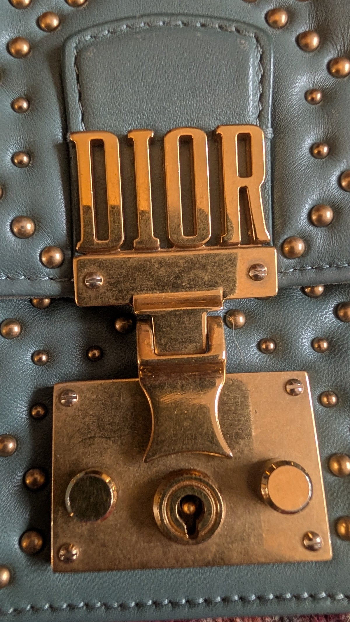 Close-up of a Dior bag featuring a large gold-tone metal clasp with the DIOR logo in raised letters, attached to a blue-gray leather flap with gold studs. Pre-Owned Christian Dior Small DiorAddict Cannage Studded Flap Bag – Light Blue Lambskin
