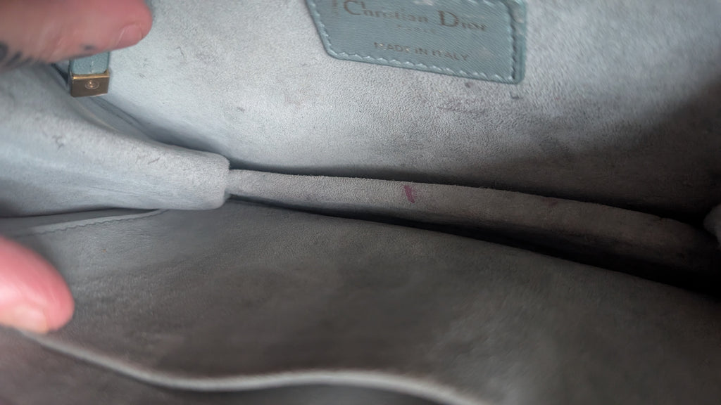 Close-up of the interior lining of a Christian Dior handbag in light gray suede or velvet, showing the brand label and a finger holding the opening. Pre-Owned Christian Dior Small DiorAddict Cannage Studded Flap Bag – Light Blue Lambskin