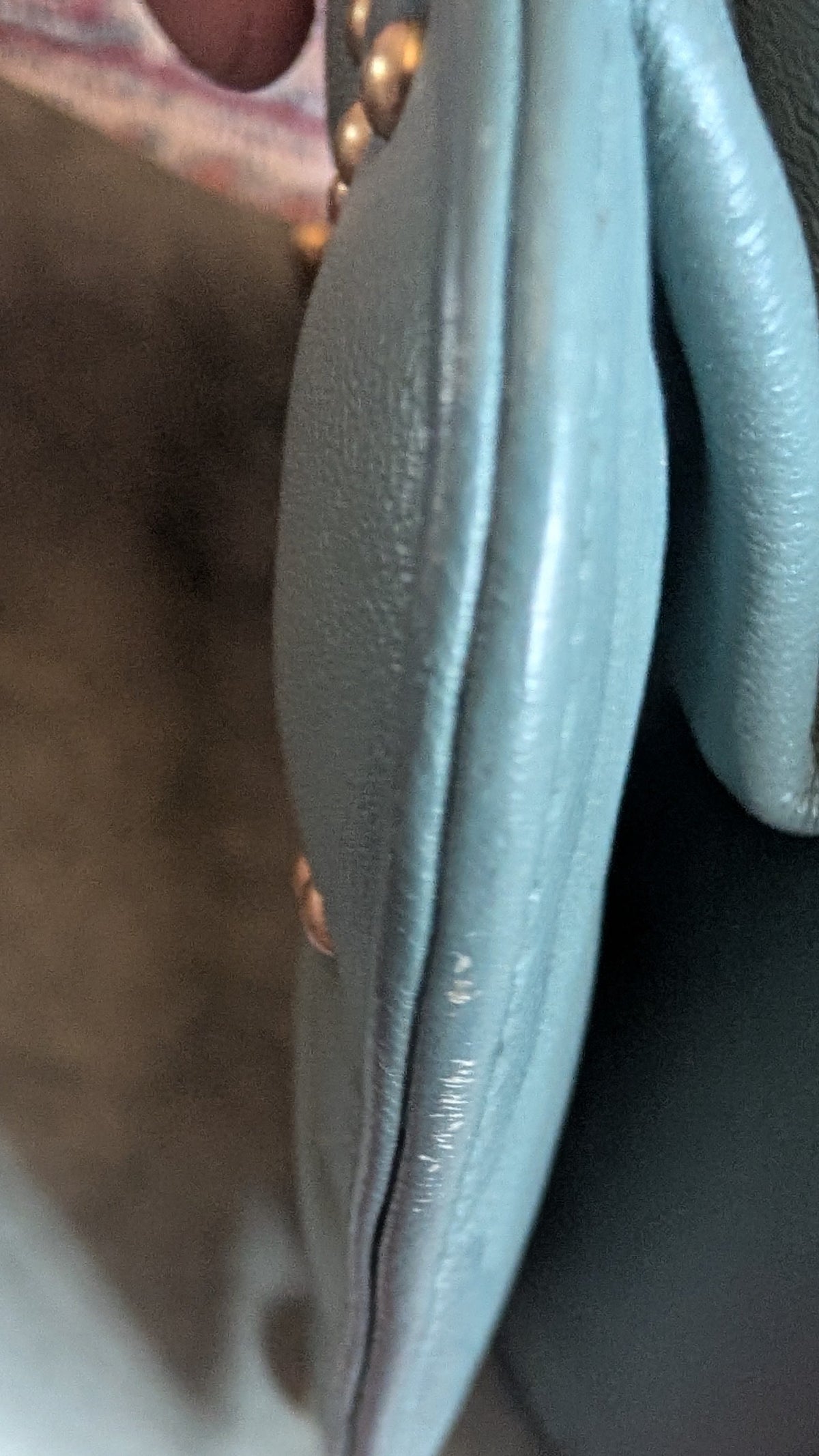 Close-up of a light blue leather clutch or wallet showing the side profile, interior lining, and a small gold-tone button detail, resting on a patterned red rug. Pre-Owned Christian Dior Small DiorAddict Cannage Studded Flap Bag – Light Blue Lambskin