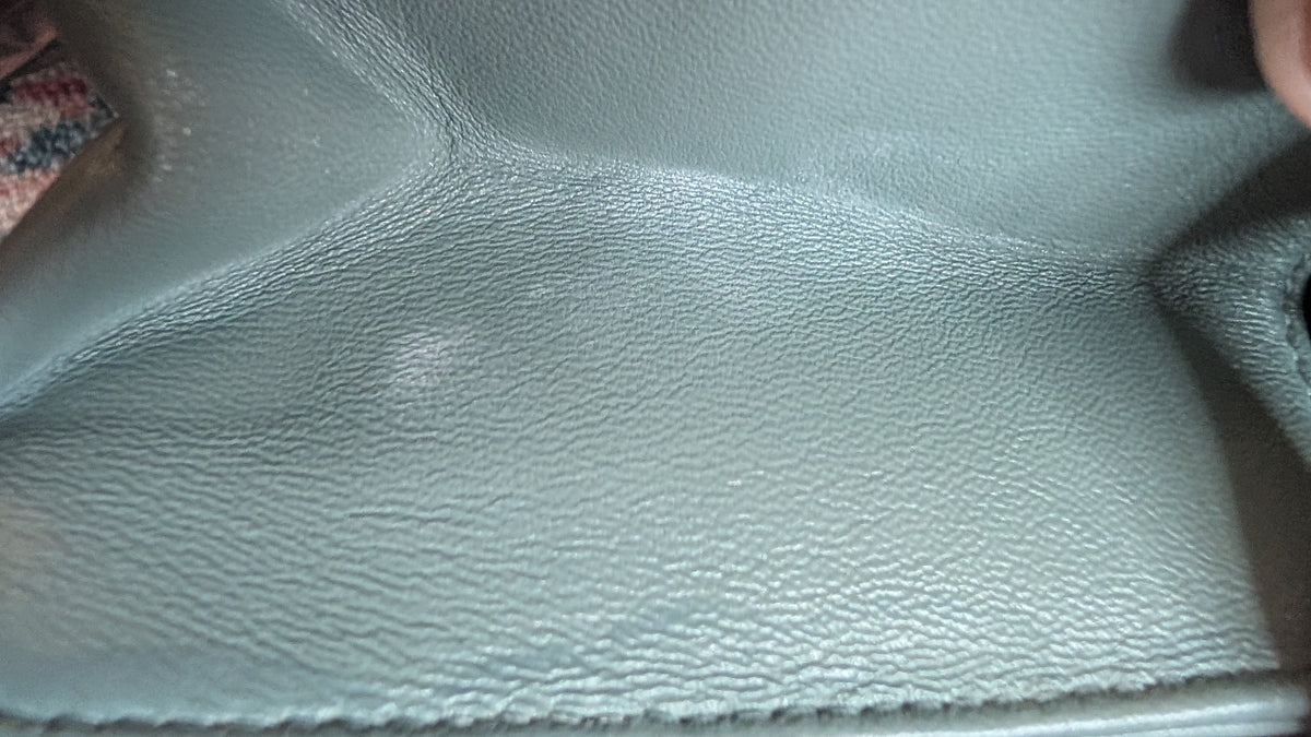 Close-up of a textured leather surface in a muted sage green color, showing the grain pattern and stitching along the bottom edge. Pre-Owned Christian Dior Small DiorAddict Cannage Studded Flap Bag – Light Blue Lambskin