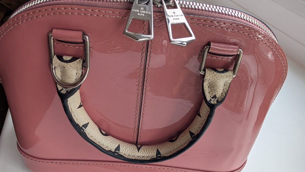 A close-up of a glossy pink patent leather handbag with silver-tone hardware and a beige-and-black patterned handle, shown on a light  surface. 