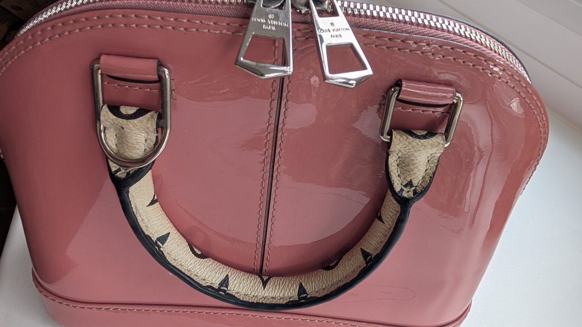 A close-up of a glossy pink patent leather handbag with silver-tone hardware and a beige-and-black patterned handle, shown on a light  surface. 