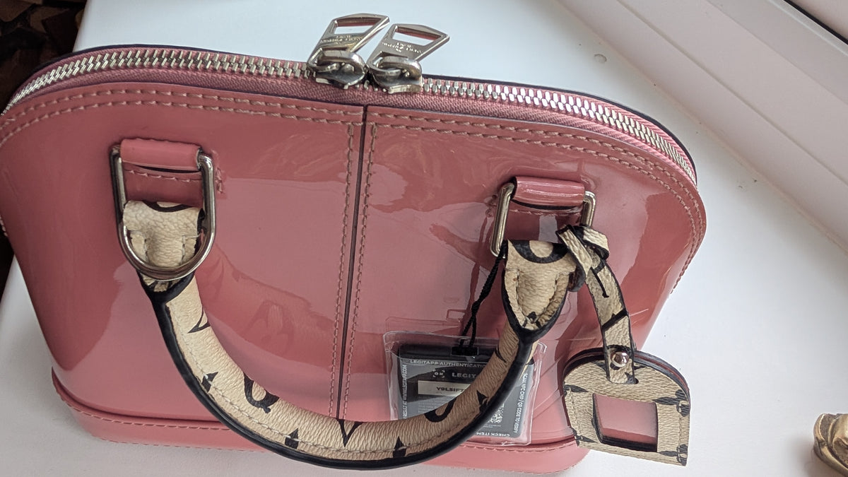 A pink patent leather cosmetic bag with a top zipper, patterned shoulder strap, and silver-tone hardware, shown on a white surface. Pre-Owned LOUIS VUITTON ALMA BB – ROSE BLUSH MONOGRAM VERNIS MIROIR