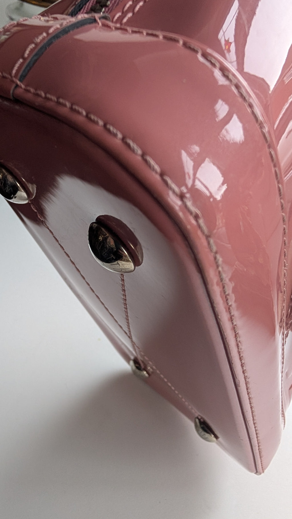 Close-up of a glossy dusty pink patent leather handbag corner, showing silver-tone metal studs and stitching on a white surface. Pre-Owned LOUIS VUITTON ALMA BB – ROSE BLUSH MONOGRAM VERNIS MIROIR