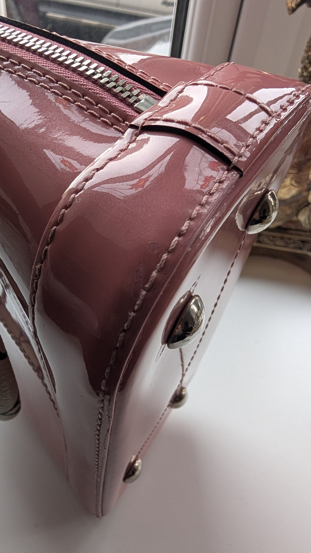 Close-up of a glossy mauve-pink patent leather handbag corner with silver-tone hardware and stitching, resting on a white surface near a window. Pre-Owned LOUIS VUITTON ALMA BB – ROSE BLUSH MONOGRAM VERNIS MIROIR