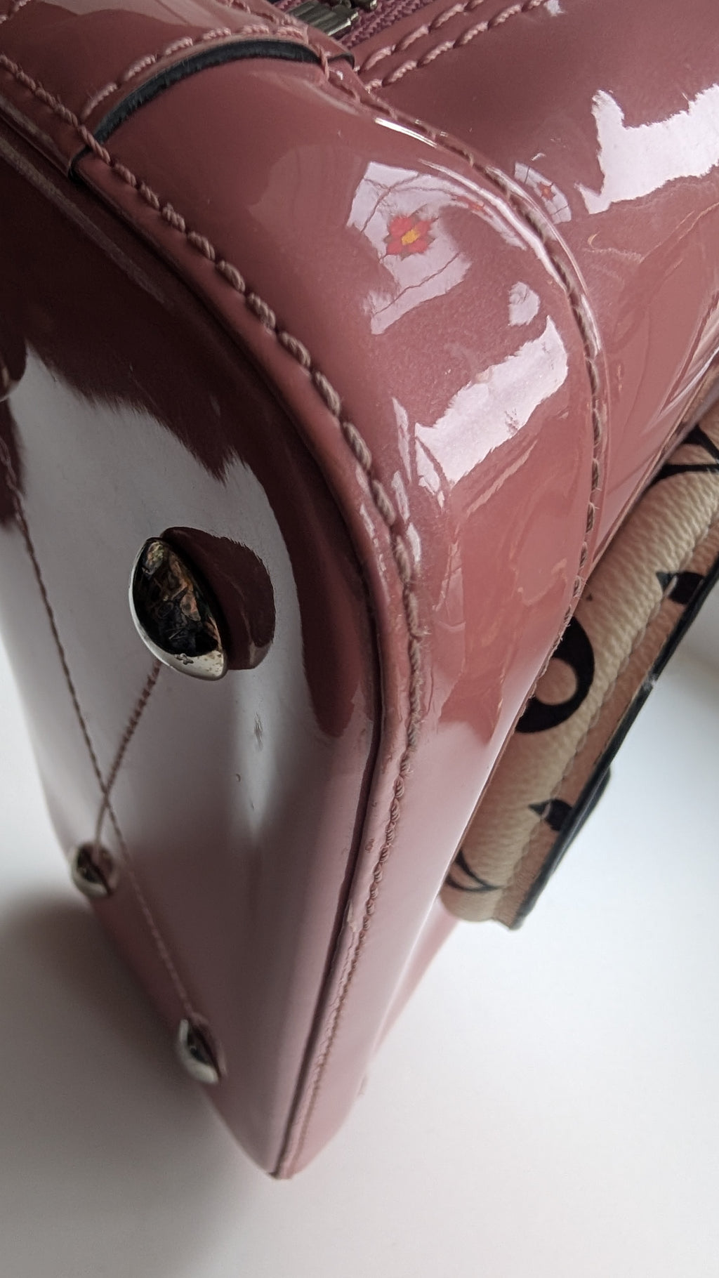 Close-up of a glossy mauve-pink patent leather handbag corner with silver-tone hardware and a patterned fabric detail visible on the right side, photographed on a white background. Pre-Owned LOUIS VUITTON ALMA BB – ROSE BLUSH MONOGRAM VERNIS MIROIR