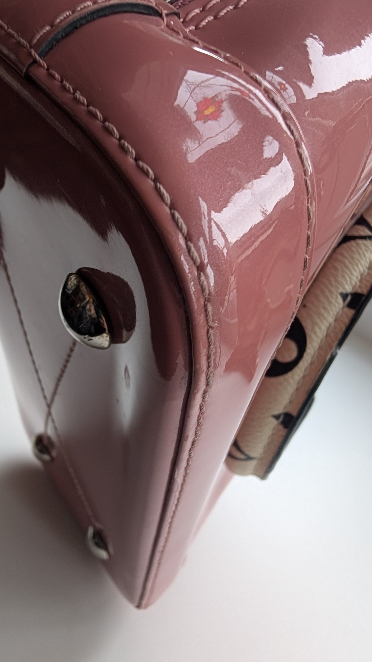 Close-up of a glossy mauve-pink patent leather handbag corner with silver-tone hardware and a patterned fabric detail visible on the right side. Pre-Owned LOUIS VUITTON ALMA BB – ROSE BLUSH MONOGRAM VERNIS MIROIR