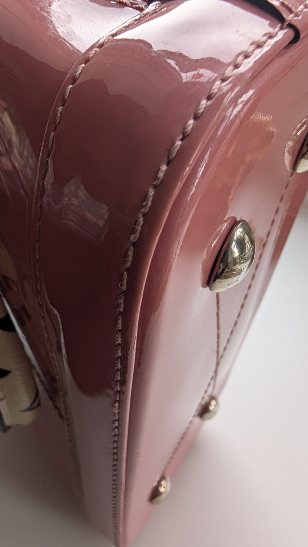 Close-up of a glossy mauve-pink patent leather bag corner with silver-tone studs and contrast stitching on a white background. 