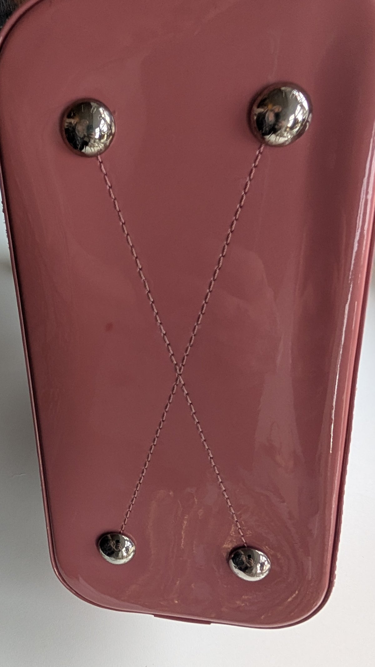 Close-up of a glossy dusty rose pink bag or case with two silver metal studs connected by thin chains, forming an X shape on the front. Pre-Owned LOUIS VUITTON ALMA BB – ROSE BLUSH MONOGRAM VERNIS MIROIR
