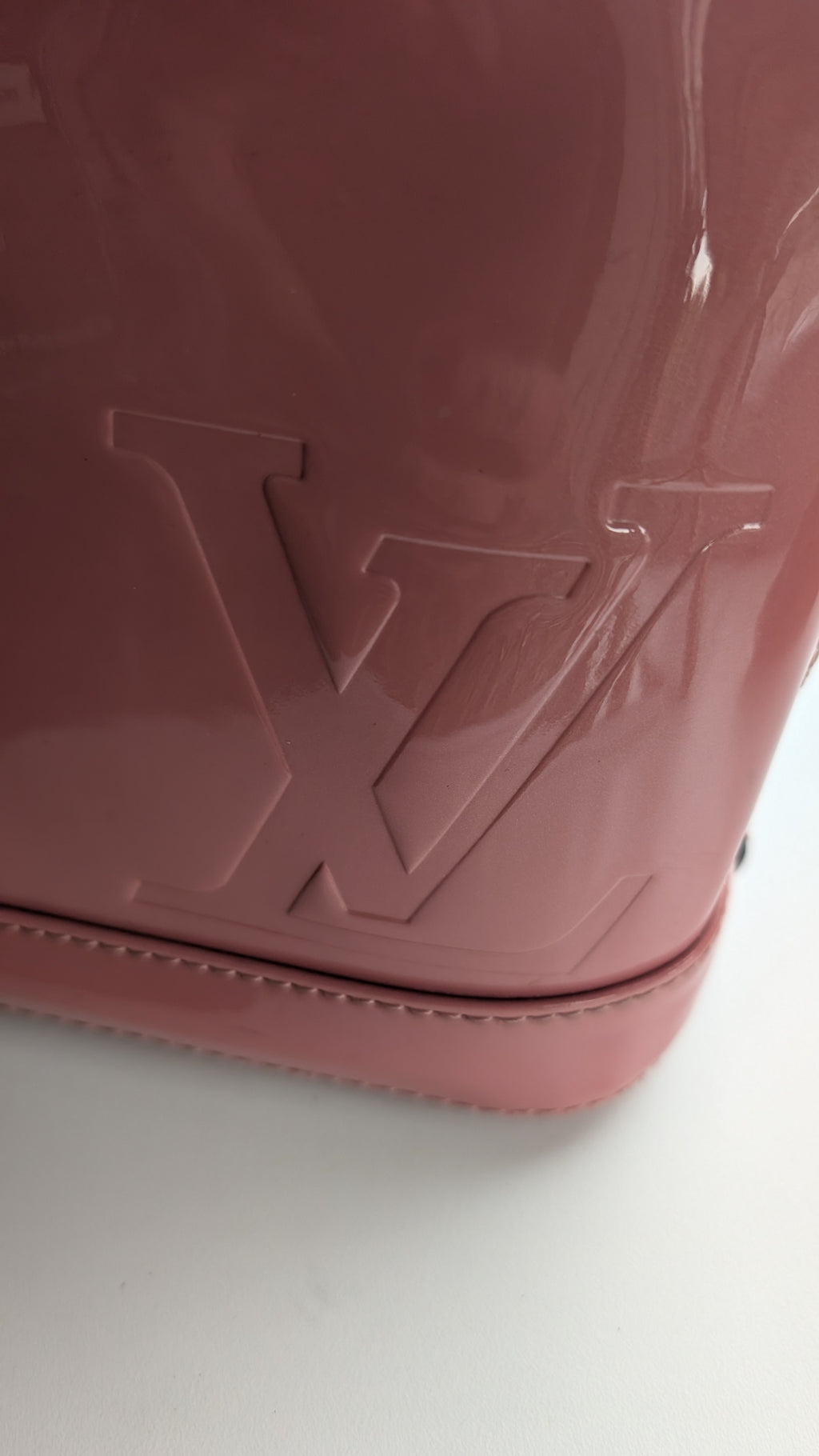 Close-up of a glossy pink Louis Vuitton handbag or case corner with an embossed LV logo and visible whipstitching along the base, photographed on a white surface. Pre-Owned LOUIS VUITTON ALMA BB – ROSE BLUSH MONOGRAM VERNIS MIROIR